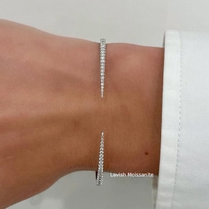 Puede incluir: Una pulsera de puño abierta plateada con una fila de pequeñas piedras transparentes. La pulsera se lleva en una muñeca junto a una camisa blanca de manga larga. El texto "Lavish Moissanite" es visible.