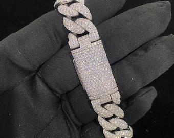 May include: A silver Cuban link chain bracelet with a clasp closure, fully encrusted with clear cubic zirconia stones. The bracelet is shown on a black surface.