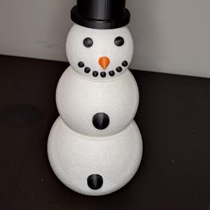 Può includere: Statuetta di pupazzo di neve stampata in 3D. Il pupazzo di neve ha un corpo bianco con cappello a cilindro nero, occhi e bottoni. Presenta un naso a carota arancione e una bocca sorridente. Decorazione festosa.