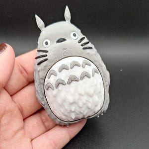 Puede incluir: Una figura impresa en 3D de Totoro, en gris y blanco, personaje de la película de animación "Mi vecino Totoro". La figura tiene una superficie texturizada, con detalles negros para los ojos, la nariz y los bigotes. El vientre presenta un patrón de formas curvas.