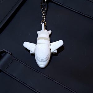 Può includere: Un portachiavi bianco a forma di aereo con una catena d'argento. Il portachiavi presenta un design dettagliato con ali e coda. Lo sfondo è una superficie nera.