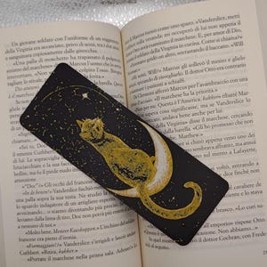 Può includere: Un segnalibro nero con una luna crescente dorata e un gatto nero con dettagli dorati. Il gatto è rannicchiato sulla luna, guardando una stella. Lo sfondo è blu scuro con stelle dorate.