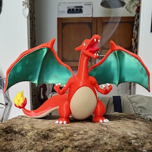 Può includere: Una statuetta rossa di Charizard con ali verdi e rosse, pancia color crema e una fiamma sulla coda. La figura del drago è in posa con la bocca aperta, che emette una voluta di fumo. È un oggetto da collezione.
