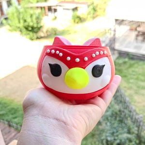 May include: A red and white owl-shaped planter with a yellow beak and black eyes. The planter is held in a hand, with a blurred background of greenery and a mountain range. The owl has white accents and a decorative design.