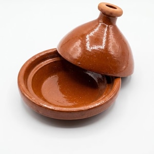 Peut inclure: Un plat à tajine en terre cuite avec une finition vernissée brun rougeâtre. L'objet se compose d'une base circulaire large et d'un couvercle conique surmonté d'un petit bouton de préhension. L'intérieur de la base est lisse et profond, tandis que l'extérieur du couvercle présente une surface brillante et réfléchissante qui met en valeur la couleur naturelle de l'argile.