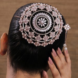 Op de afbeelding: Een sierlijke, cirkelvormige haaraccessoire met een bloemmotief. Het stuk heeft een centraal bloemdesign, omringd door kleinere bloemen, parels en kristallen. Een delicate ketting met een parel hangt aan de zijkant. De accessoire is geplaatst op donker haar.