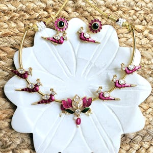 Puede incluir: Un conjunto de collar y pendientes dorados con un diseño de pavo real con esmalte rosa y detalles de piedras preciosas transparentes. El collar tiene un colgante de flor de loto con una cuenta roja. Los pendientes tienen forma de flor con un centro negro.