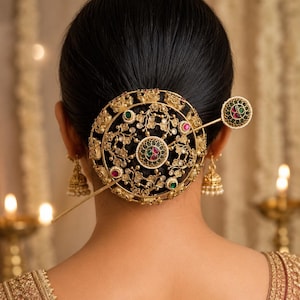 May include: A gold-toned circular hair accessory featuring intricate filigree metalwork, embedded with small green and red stones, secured by a matching decorative pin. The piece is worn over a dark hair bun, complemented by gold-toned bell-shaped earrings.