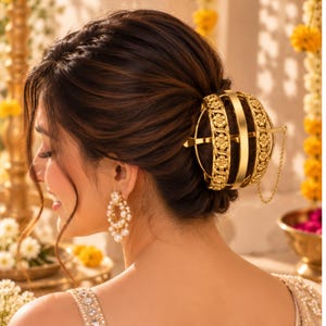 Puede incluir: Un accesorio para el cabello esférico de tono dorado con grabados florales intrincados y una banda central, asegurado con un pasador horizontal y una cadena fina. Se lleva en un recogido oscuro, junto a pendientes de perlas colgantes.
