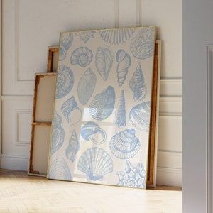 May include: A framed print with a white background and a blue pattern of various seashells. The print is in a gold frame and is leaning against a white wall.