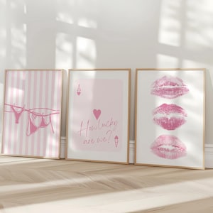 May include: Three framed prints with a light wood finish lean against a wall. The prints feature pink and white designs: striped background with lingerie, a playing card with text, and three lip prints. The text on the card reads "How lucky are we?"