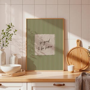 May include: A framed print with a green striped background and a square patch with the text "so good to be home." The print is displayed on a wooden countertop with a white bowl, a wooden cutting board, and a small white cup.