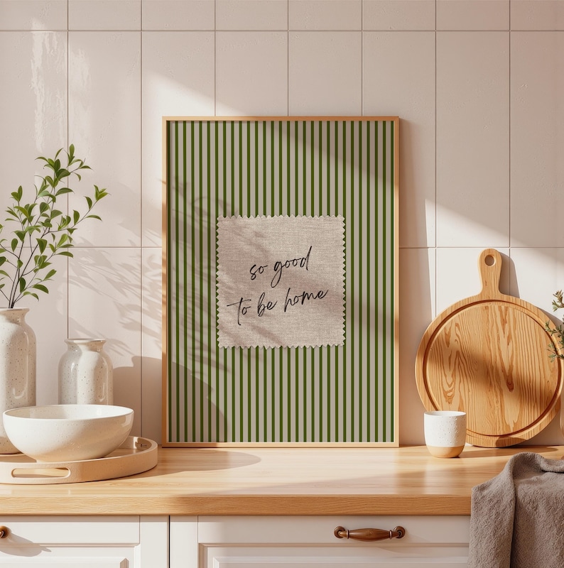 May include: A framed print with a green striped background and a square patch with the text "so good to be home." The print is displayed on a wooden countertop with a white bowl, a wooden cutting board, and a small white cup.