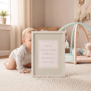 May include: A framed print with a light wood frame and a striped background. The print has the text "YOU'LL NEVER KNOW little one HOW MUCH I LOVE YOU" in a decorative font. A baby is in the background.