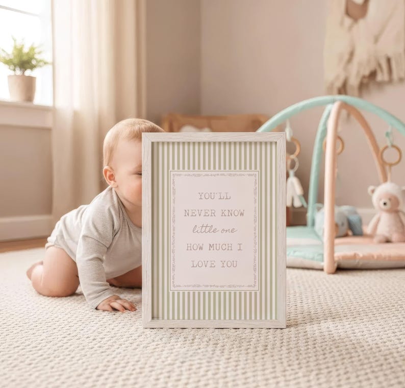 May include: A framed print with a light wood frame and a striped background. The print has the text "YOU'LL NEVER KNOW little one HOW MUCH I LOVE YOU" in a decorative font. A baby is in the background.