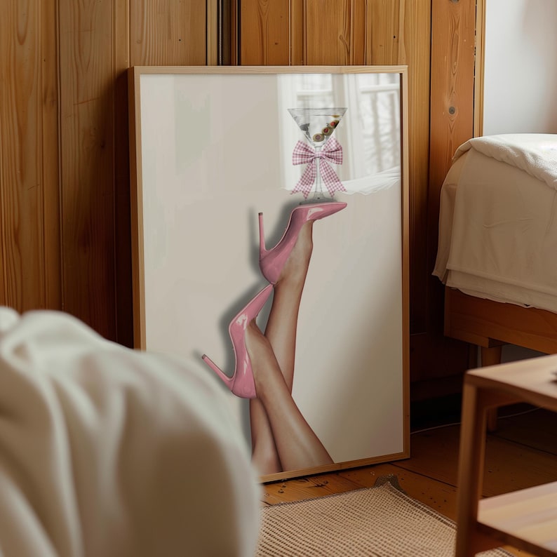 May include: A framed print featuring a martini glass with a pink gingham bow and pink high heels. The heels are positioned with the legs crossed, and the martini glass is balanced on the feet. The artwork has a light pink background.