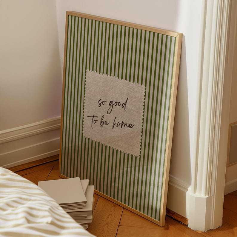 May include: A framed print with a green and white striped background and a linen patch in the center. The linen patch has the text "so good to be home" in a handwritten style. The frame is a light wood color.
