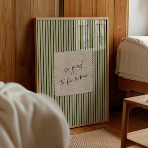 May include: A framed wall art piece with a green and white striped background. A square patch of fabric with the words "so good to be home" is centered on the artwork. The frame is a light wood color.