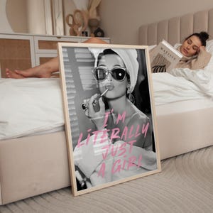 May include: Black and white framed print of a woman applying lipstick, wearing sunglasses and a towel. The pink text reads "I'M LITERALLY JUST A GIRL". The print is propped against a bed.
