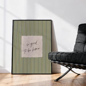 May include: A framed print with a green and white striped background and a square patch with the text "so good to be home." The print is displayed on a wooden floor next to a black chair.