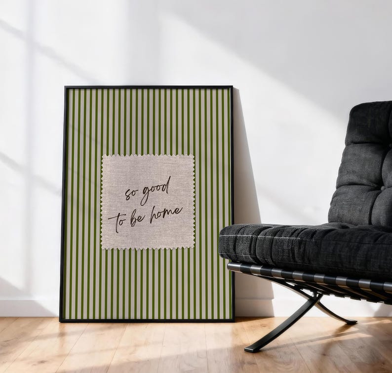 May include: A framed print with a green and white striped background and a square patch with the text "so good to be home." The print is displayed on a wooden floor next to a black chair.