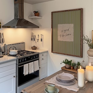 May include: A kitchen scene featuring a stainless steel stove and range hood. A framed artwork with green and white stripes and the text "so good to be home" hangs on the wall. A wooden table is set with plates, a cup, and candles.