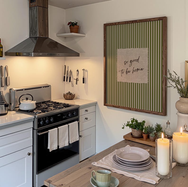 May include: A kitchen scene featuring a stainless steel stove and range hood. A framed artwork with green and white stripes and the text "so good to be home" hangs on the wall. A wooden table is set with plates, a cup, and candles.