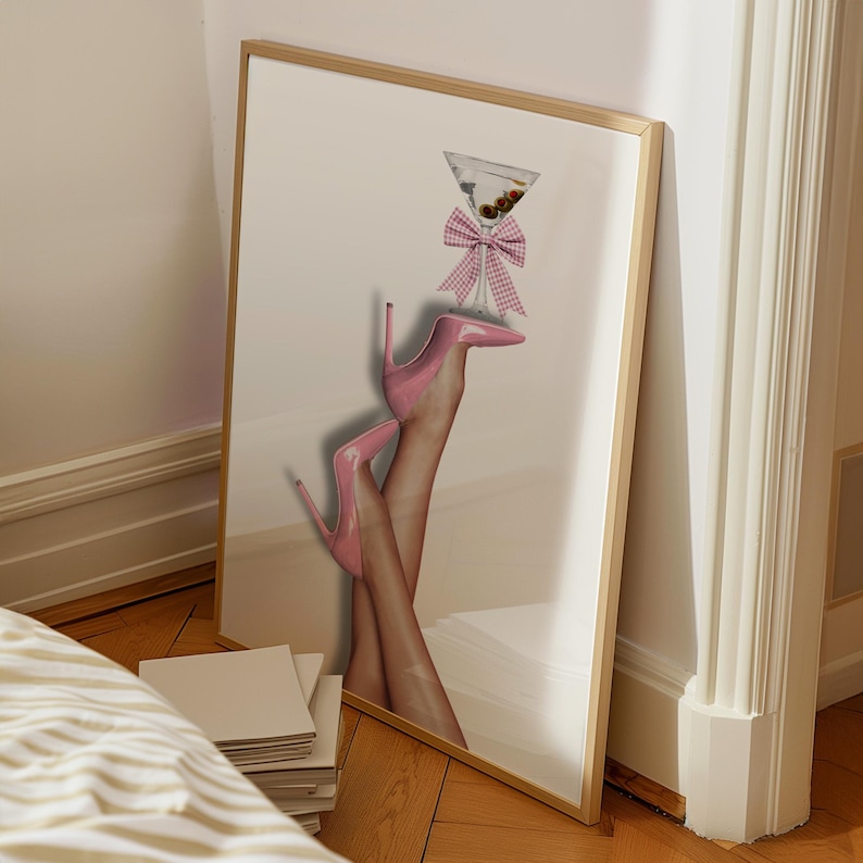 May include: A framed wall art print featuring a pair of legs wearing glossy pink high-heeled pumps. A martini glass with olives is balanced on the toe of one shoe, decorated with a pink and white gingham bow tied around the stem of the glass.