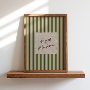 Op de afbeelding: Een ingelijste print met een houten frame op een houten plank. De print heeft een groen-witte gestreepte achtergrond met een vierkante, getextureerde patch in het midden. De patch heeft de woorden "so good to be home" in cursief.