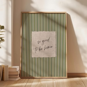 May include: A framed print with a green and white striped background. A square patch with the text "so good to be home" is centered on the print. The frame is a light brown color.