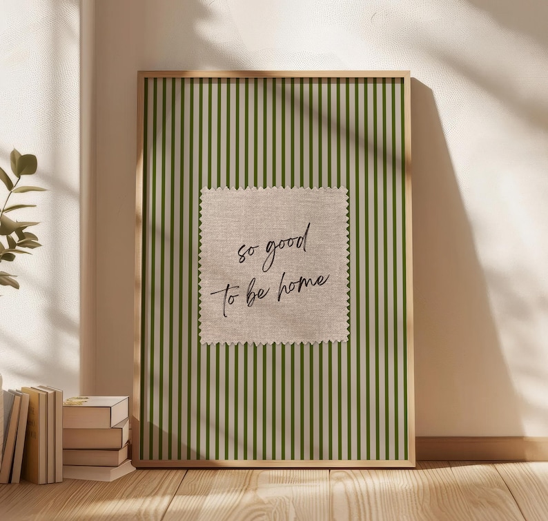 May include: A framed print with a green and white striped background. A square patch with the text "so good to be home" is centered on the print. The frame is a light brown color.