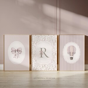 May include: Three framed nursery art prints. The prints feature a bow, the letter "R", and a hot air balloon. The color palette includes soft pinks, whites, and grays. The prints are in gold frames and are displayed on a wooden floor.