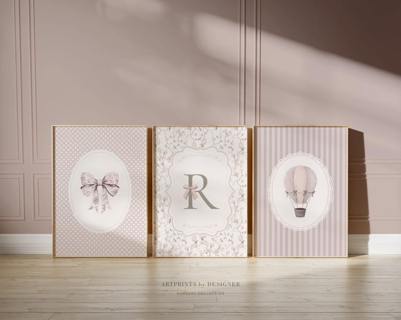 May include: Three framed nursery art prints. The prints feature a bow, the letter "R", and a hot air balloon. The color palette includes soft pinks, whites, and grays. The prints are in gold frames and are displayed on a wooden floor.
