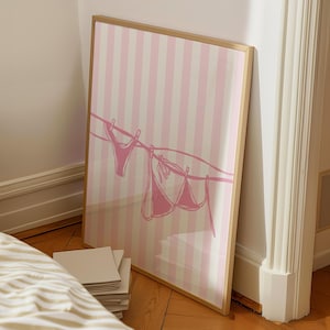 May include: A framed art print featuring a pink and white striped background. A line with a pink bikini top and bottom is drawn across the image. The frame is a light brown color.