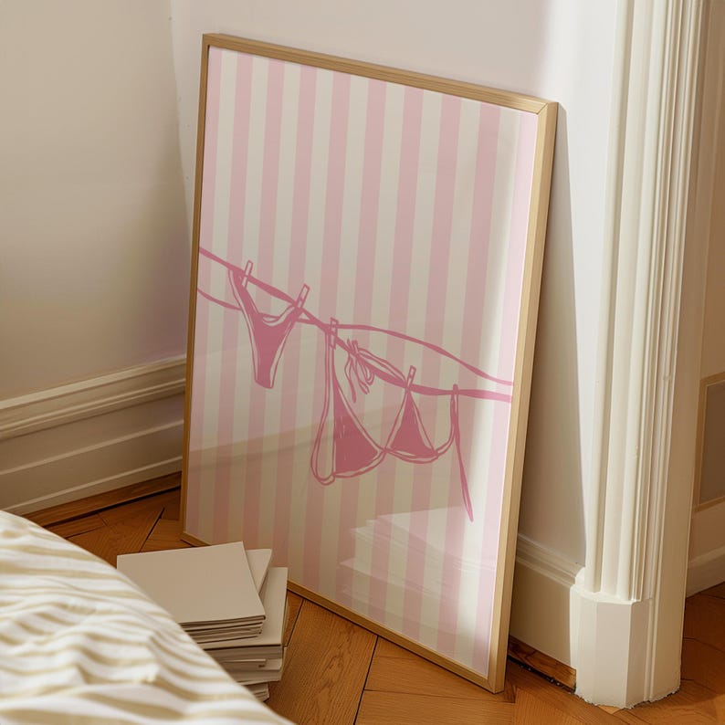 May include: A framed art print featuring a pink and white striped background. A line with a pink bikini top and bottom is drawn across the image. The frame is a light brown color.