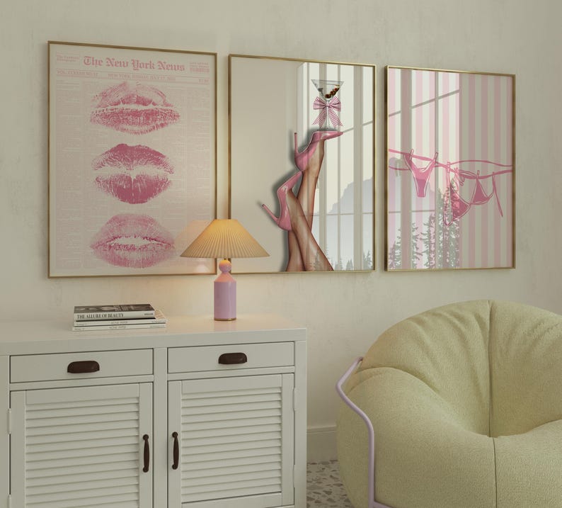 May include: Three framed art prints with gold frames. One print features pink lipstick prints, another shows legs in pink heels holding a martini, and the third depicts pink lingerie hanging on a clothesline. A pink lamp sits on a white cabinet.