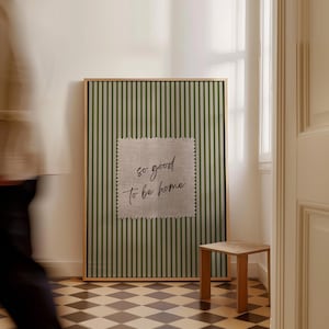 May include: A framed wall art piece with a green and white striped background. A square patch of fabric with the text "so good to be home" is centered on the artwork. A small wooden side table is next to the artwork.