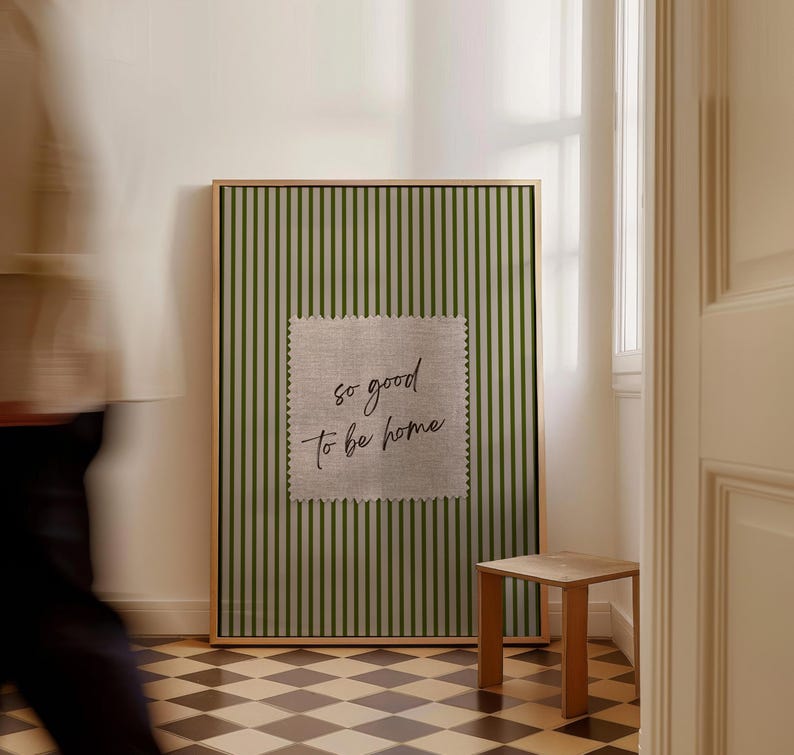 May include: A framed wall art piece with a green and white striped background. A square patch of fabric with the text "so good to be home" is centered on the artwork. A small wooden side table is next to the artwork.