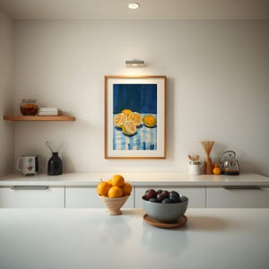 May include: A framed painting of lemons on a blue and white checkered tablecloth. The painting is hanging on a white wall above a white countertop with a bowl of lemons and a bowl of plums.