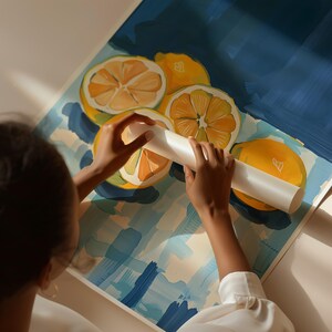 May include: A person is rolling out a piece of paper with a white roller. The paper has a blue and white checkered background with a painting of lemons on it. The lemons are yellow and have a white background.