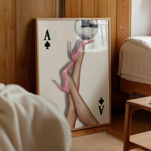 May include: Framed art print showing legs in pink high heels balancing a disco ball. The print includes the Ace of Spades card symbol and text. The artwork is displayed in a room with wooden paneling.