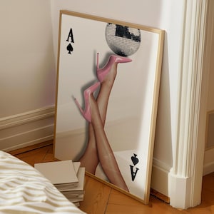 May include: A framed art print featuring a pair of legs wearing pink high heels, balancing a disco ball. The artwork is designed to resemble a playing card, with the Ace of Spades symbols.