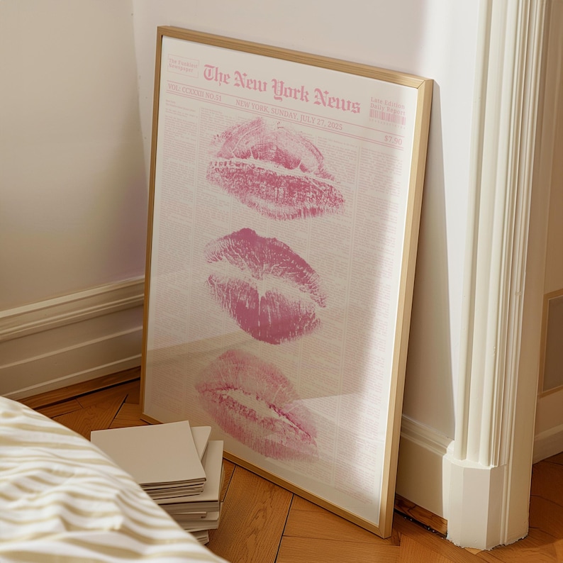 May include: A framed print featuring three pink lip imprints on a newspaper background. The newspaper is titled "The New York News" and dated July 27, 2015. The frame is a light brown color.