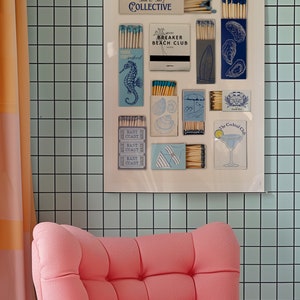 May include: A white framed poster with a collection of matchboxes featuring various designs, including seashells, a cocktail glass, and a surfboard. The poster is titled "Sun & Surf Collective" and is displayed on a light blue tiled wall.