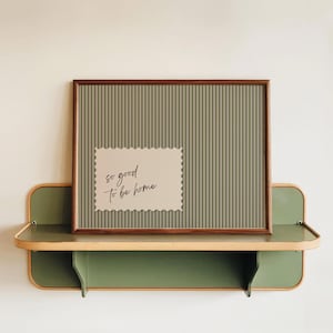 Puede incluir: Una obra de arte enmarcada con un fondo verde a rayas y una tarjeta más pequeña con bordes festoneados que dice "so good to be home". La obra de arte se exhibe en una estantería verde y beige, creando una estética acogedora.
