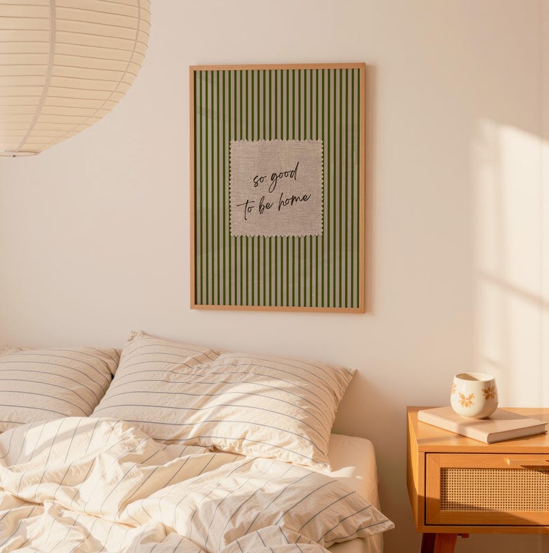 May include: A framed wall art piece with a green striped background and a square, textured patch in the center. The patch features the handwritten phrase "so good to be home" in black script. The frame is a light brown color.
