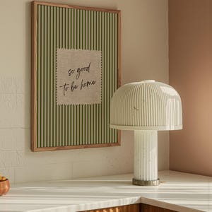 May include: A framed artwork with green vertical stripes and a linen patch that reads "so good to be home." A white lamp with a ribbed shade and base sits on a white countertop. The lamp has a cylindrical base and a rounded top.