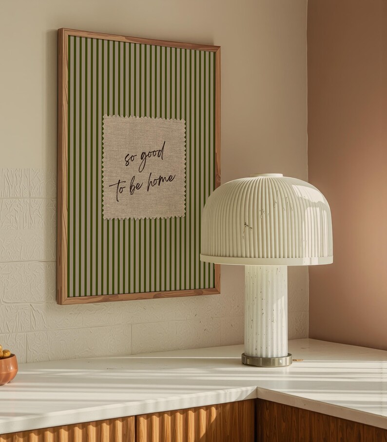 May include: A framed artwork with green vertical stripes and a linen patch that reads "so good to be home." A white lamp with a ribbed shade and base sits on a white countertop. The lamp has a cylindrical base and a rounded top.