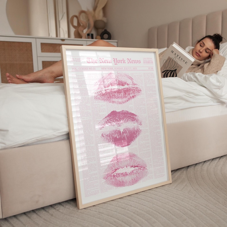 May include: A framed vertical poster featuring a light pink background with the text The New York News at the top. The design includes three distinct pink lipstick kiss marks stacked vertically in the centre, overlaid on a newspaper-style layout.
