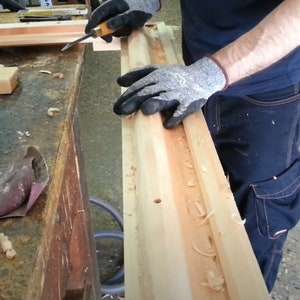 Puede incluir: Una persona con guantes de trabajo utiliza un cincel para tallar una ranura en una pieza de madera. La madera est&aacute; sobre un banco de trabajo con otras herramientas de carpinter&iacute;a.